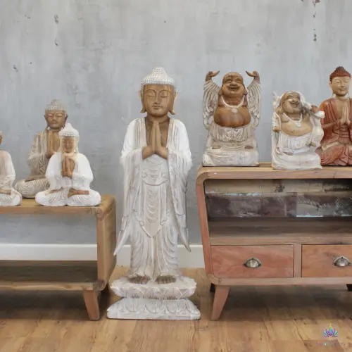 Grande Statue Bouddha Debout en Bois Blanchi – 100 cm, Mains Jointes 3 Statue de Bouddha debout en bois blanchi mise en scène devant un mur en béton, entre des Bouddhas assis