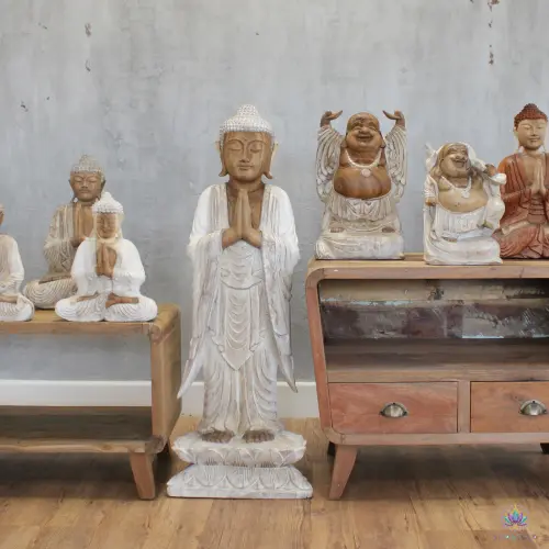 Grande Statue Bouddha Debout en Bois Blanchi – 100 cm, Mains Jointes 2 Statue de Bouddha debout en bois blanchi entre deux meubles en bois, entourée d'autres statues