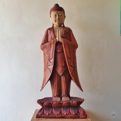 Grande Statue Bouddha Debout en Bois de Suar – 100 cm, Mains Jointes 2 Statue de Bouddha debout sculptée à la main, yeux clos, posée sur un socle en bois devant un mur blanc
